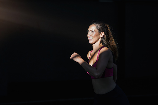 Young woman with fit body running against dark background. Female model in sportswear exercising outdoors.