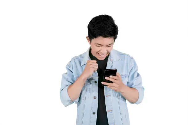 Portrait of a young Asian man looks happy while reading a good news on the smartphone, isolated on white background
