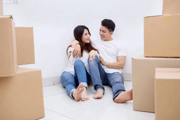 Image of happy Asian couple sitting with a stack of cardboard boxes in the new house