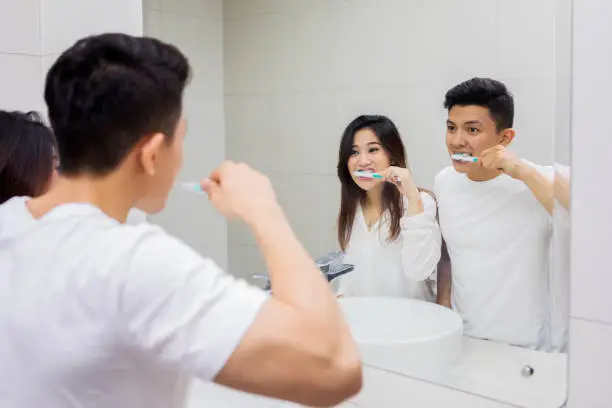 Picture of Asian couple brushing teeth in the bathroom while looking at mirror