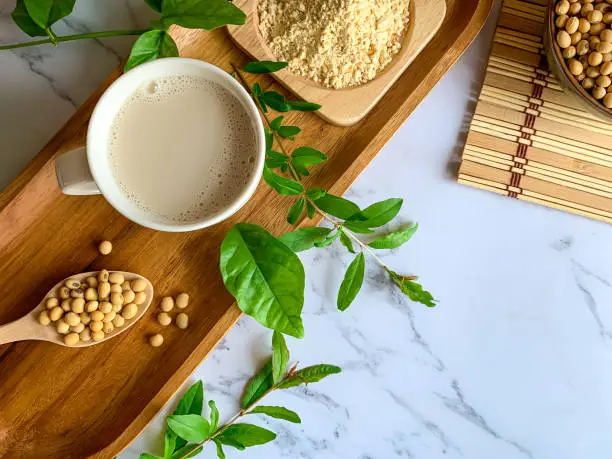 Top view of soymilk cups,soybean seeds on wooden spoon and soy milk powder in a wooden plate background white marble table, is a healthy drink for women because of the "isoflavones" substance, making it clear and helping to slow down aging