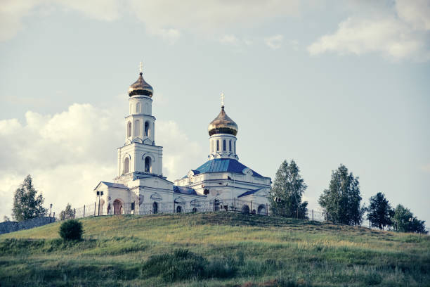 A golden domed bell tower is framed by leafy tree branches, set against a dramatic cloudy sky, highlighting the intricate architecture.