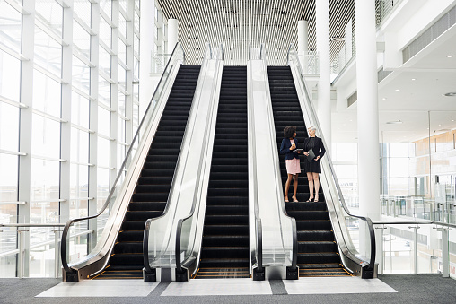 Full length shot of two businesswomen having a discussion while coming down an escalator in the workplace