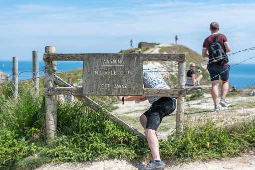 Lulworth, Dorset, UK - July 6, 2019: Young men ignore a keep out sign and venture on to a piece of cliff edge on the Dorset coast.