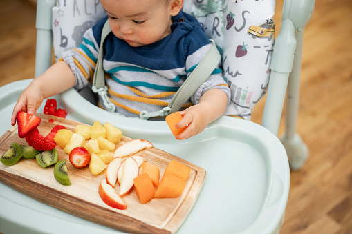 Close-up of a baby boy sitting in a high chair at home. He is exploring different fruits from a wooden try in front of him.