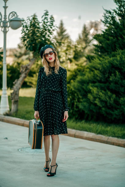 jeune femme dans la robe noire de point de polka de cru avec la valise rétro dans la main posant à l'extérieur - polka dot suitcase retro revival women photos et images de collection