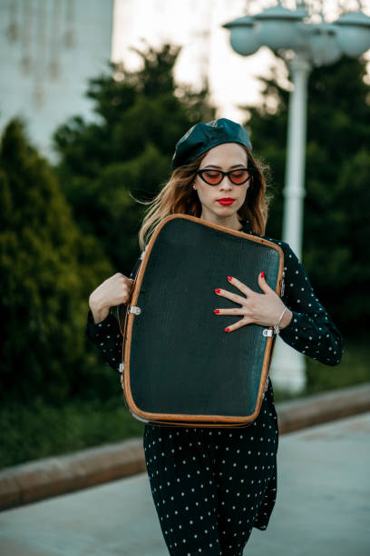 jeune femme dans la robe noire de point de polka de cru avec la valise rétro dans la main posant à l'extérieur - polka dot suitcase retro revival women photos et images de collection