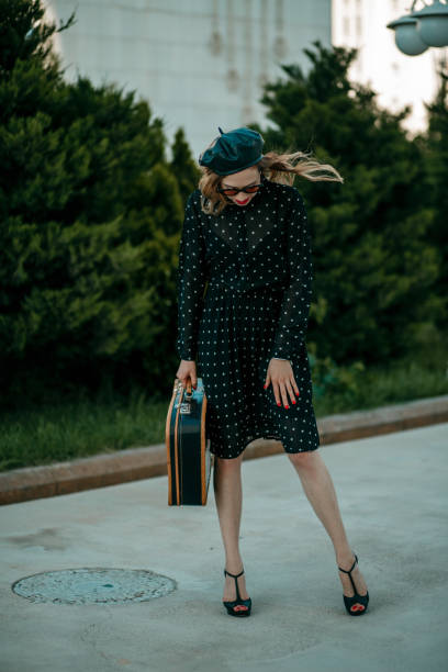 jeune femme dans la robe noire de point de polka de cru avec la valise rétro dans la main posant à l'extérieur - polka dot suitcase retro revival women photos et images de collection