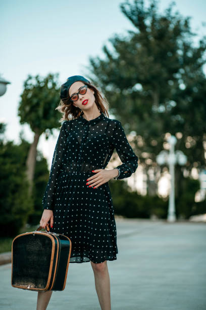 jeune femme dans la robe noire de point de polka de cru avec la valise rétro dans la main posant à l'extérieur - polka dot suitcase retro revival women photos et images de collection