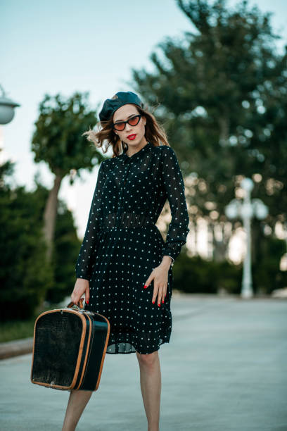 jeune femme dans la robe noire de point de polka de cru avec la valise rétro dans la main posant à l'extérieur - polka dot suitcase retro revival women photos et images de collection