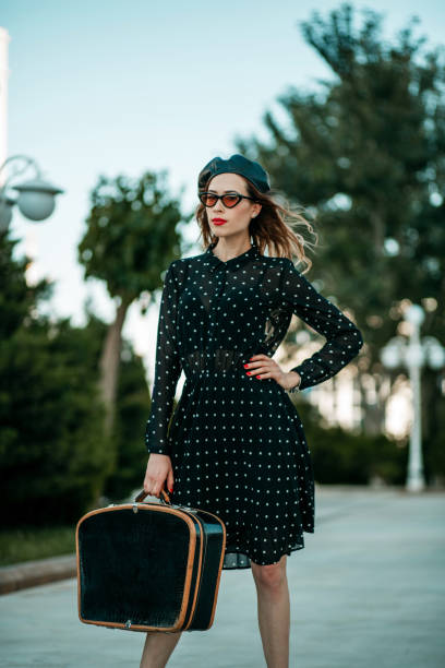 jeune femme dans la robe noire de point de polka de cru avec la valise rétro dans la main posant à l'extérieur - polka dot suitcase retro revival women photos et images de collection