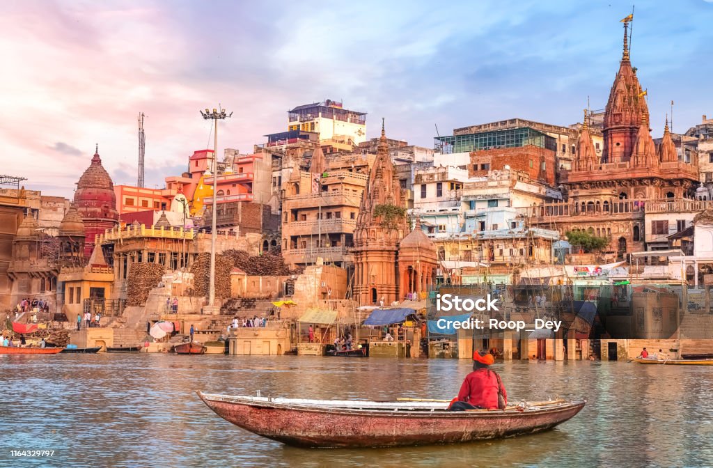 Hindu sadhu sitting on a boat overlooking Varanasi city architecture at sunset Ancient Varanasi city architecture and Ganges river ghat at sunset with view of an Indian sadhu sitting a boat on the river Ganges. Varanasi Stock Photo