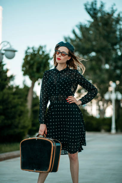 jeune femme dans la robe noire de point de polka de cru avec la valise rétro dans la main posant à l'extérieur - polka dot suitcase retro revival women photos et images de collection