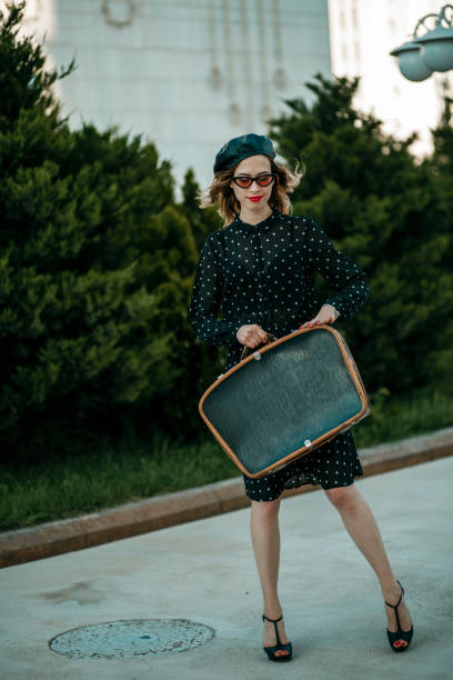 jeune femme dans la robe noire de point de polka de cru avec la valise rétro dans la main posant à l'extérieur - polka dot suitcase retro revival women photos et images de collection