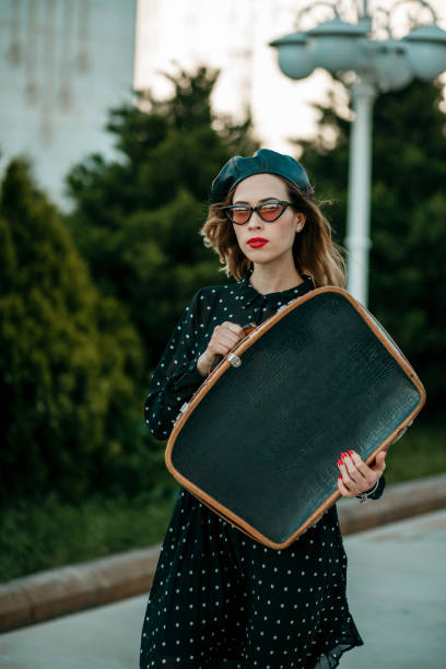 jeune femme dans la robe noire de point de polka de cru avec la valise rétro dans la main posant à l'extérieur - polka dot suitcase retro revival women photos et images de collection