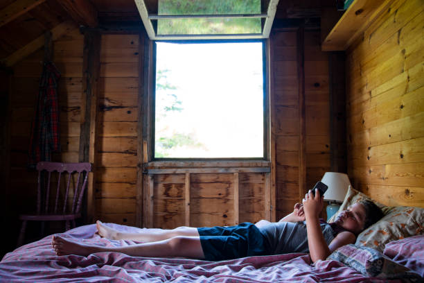 夏の小屋でリラックス。 十代前の少年は素朴な夏の小屋やコテージの寝室でスマートフォンでベッドでリラックスします。 - cabin indoors rustic bedroom ストックフォトと画像