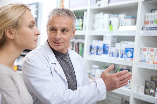 Senior pharmacist helping his female customer choosing prescription drugs from the shelf. Young woman shopping at drugstore, taking advice from mature chemist. Consumerism, friendly staff concept