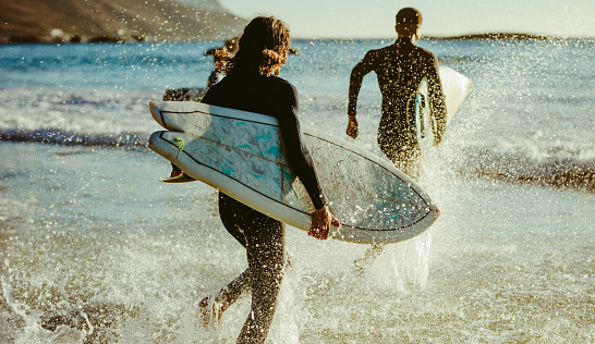 Rear view of two men going in to the sea with surfboards. Male surfers running into ocean water for surfing.