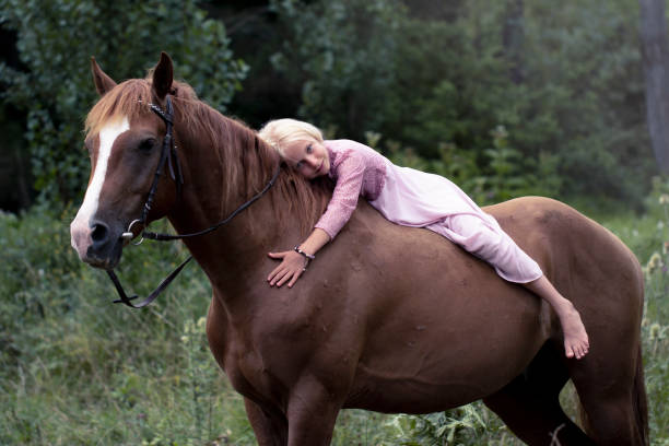 блондинка девушка с лошадью в лесу. красивая кавказская девушка с длинными светлыми волосами в розовом платье лежит на коричневой лошади и  - bareback стоковые фото и изображения