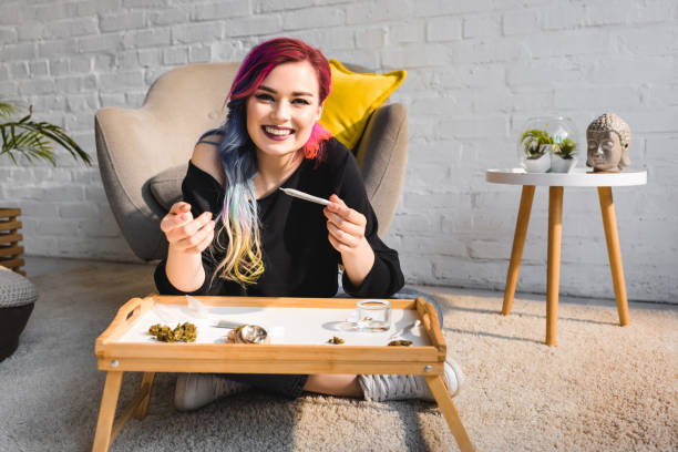 istock attractive hipster girl sitting on floor behind table, rolling joint and smiling at camera 1164348981