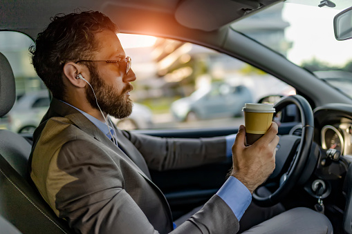 Handsome Elegant Bearded Mid Adult Businessman in Full Suit Drinking Coffee to go While Driving a Car. Businessman Manager Driving Car While Drinking a Cup of Coffee