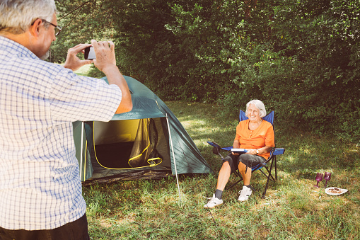 Senior couple having fun photographing in front of tent at camping place with mobile phone.