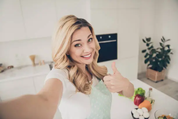 Self-portrait of her she nice attractive lovely sweet charming cute cheerful, cheery content wavy-haired lady showing thumbup in light white interior kitchen indoors