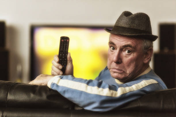 istock Senior man wearing hat holds remote control as he watches television 1164366098