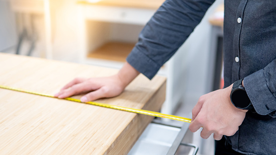 Male hand interior designer using tape measure for measuring size of wooden countertop in modern kitchen showroom in furniture store. Shopping material design for home improvement.