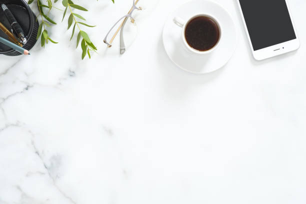 istock Flat lay, top view office table desk. Feminine desk workspace frame with coffee cup, glasses, green floral branch, smartphone and stationery on white marble background. 1164372099