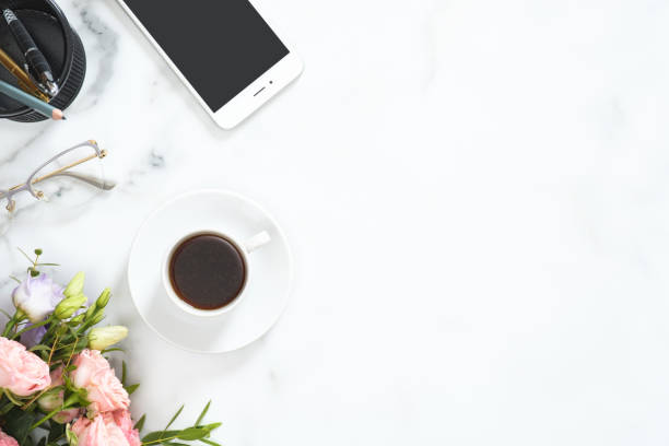 istock Flat lay, top view office table desk. Feminine desk workspace frame with coffee cup, floral bouquet, glasses, smartphone and stationery on white marble background. 1164372101