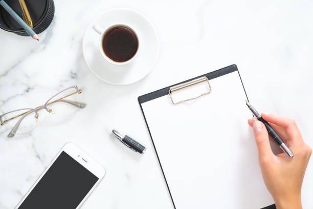 istock Woman hand write a message in blank clipboard over modern home office workspace with coffee cup, stationery and accessories on white marble background. Flat lay, top view. 1164372104