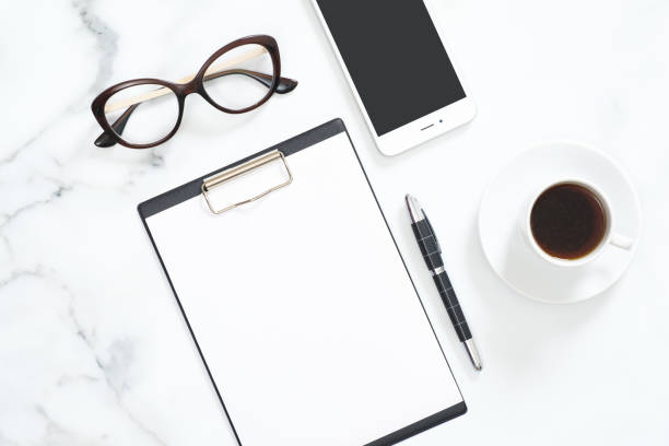 istock Modern home office desk workspace with blank paper clipboard, glasses, stationery, smartphone on white marble background. Flatlay, top view. 1164372105