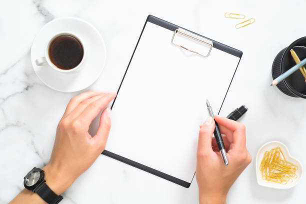 istock Woman hands holding clipboard and write a message over modern home office workspace with coffee cup, gold stationery and accessories on white marble background. Flat lay, top view. 1164372122