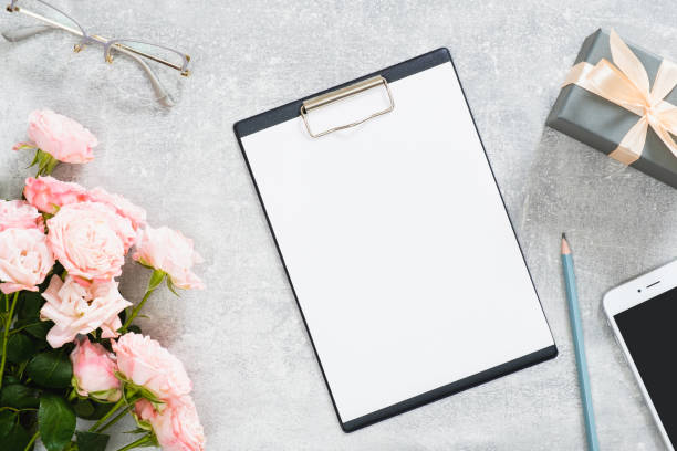 istock Modern home office workspace with paper clipboard mockup, rose flowers, gift box, glasses, mobile phone on concrete stone background. Flat lay, top view. 1164372147