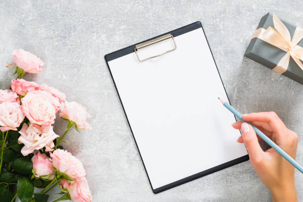 istock Home office desk workspace with rose flowers, gift box, woman hand write a message in paper clipboard on concrete stone background. Flat lay, top view modern fashion blog template. 1164372155