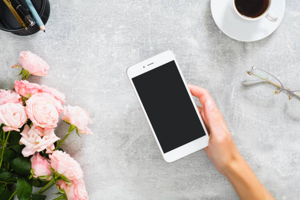 istock Female hand holding smartphone with blank screen mockup. Composition with roses flowers, coffee cup, stationery, glasses on concrete stone background. Feminine desk, beauty technology concept. 1164372157