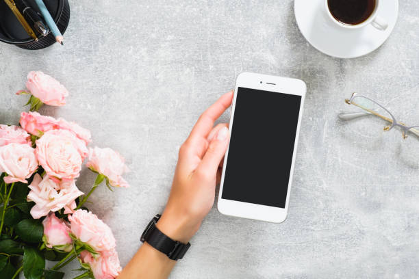 istock Female hand holding smartphone with blank screen mockup. Composition with roses flowers, coffee cup, stationery, glasses on concrete stone background. Feminine desk, beauty technology concept. 1164372160