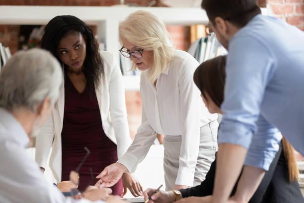 Serious mature business woman supervisor teaching workers at group meeting Serious mature woman supervisor teaching young staff workers explain new project plan at group meeting, focused old female teacher mentor training diverse business team at corporate office briefing multi generation family stock pictures, royalty-free photos & images