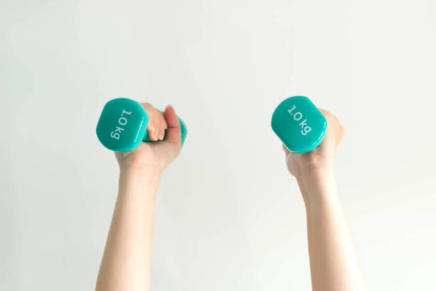 Close-up of two hands of woman lifting pastel-blue dumbbells overhead weighing 1 kilogram in each side; on white background. health care and exercise concept.(soft focus)