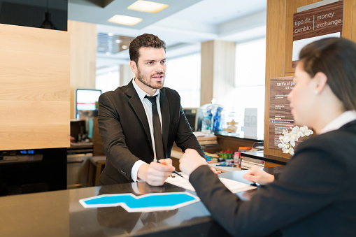 Confident handsome receptionist explaining form while assisting businesswoman at front desk in hotel