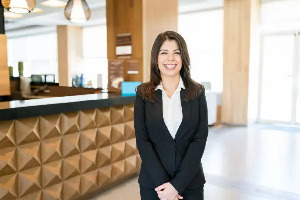 Portrait of smiling Hispanic female receptionist wearing black suit while standing in lobby at hotel