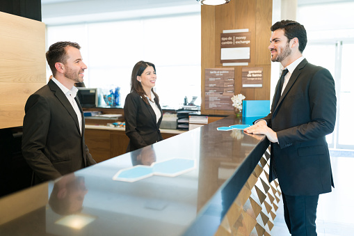 Attractive businessman talking with manager while making reservation at reception desk in lobby