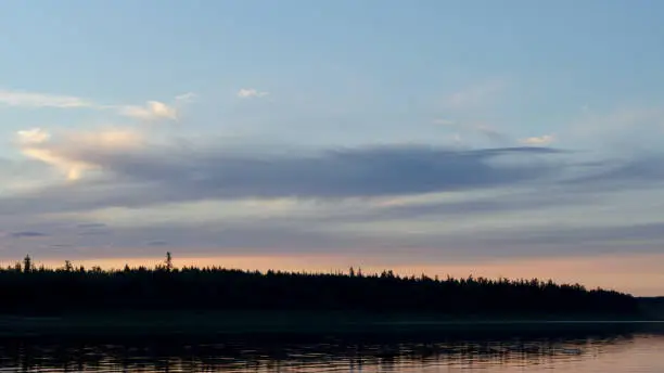 Photo of Bright blue sunset on the banks of the Northern river Viluy under the sky with clouds and a silhouette of spruce taiga in Yakutia with ripples from the wind.