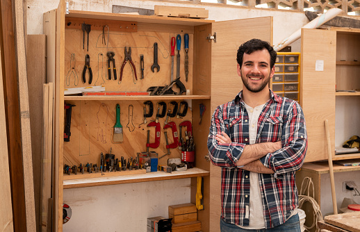 Portrait of a happy carpenter next to his tool cabinet at the workshop and looking at the camera smiling