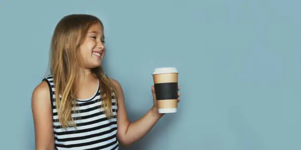 Smiling girl giving, showing coffeecup closeup side view. Drinking hot, energizing coffee to go, take away drink in disposable paper cup . Everyday routine with your child. Safe, recyclable materials