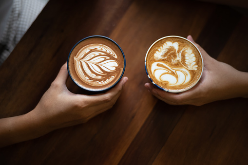 Couple hands holding two cups of coffee lattes with coffee art on top of wooden table