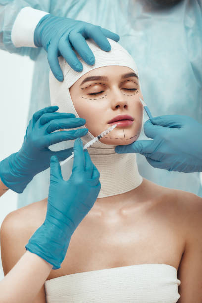 nessuna possibilità di rughe. giovane donna con la testa in bende che tengono gli occhi chiusi mentre i chirurghi plastici con guanti blu stanno lavorando con il suo viso. medici che tengono bisturi e siringa vicino al viso del paziente - plastic surgery bandage surgery women foto e immagini stock