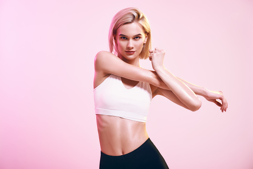 Warming up. Sporty and cute slim woman in sportswear stretching her arms and looking at camera while standing against pink background in studio. Sport. Active lifestyle. Workout