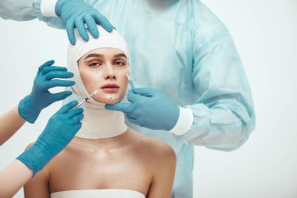 smettila di invecchiare. giovane donna attraente con la testa in bende che guarda la telecamera mentre i medici fanno un'operazione di chirurgia plastica. chirurghi plastici con guanti blu con bisturi e siringa - plastic surgery bandage surgery women foto e immagini stock